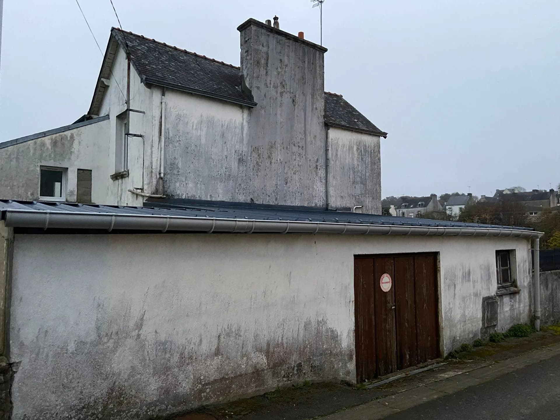 Façade enduite tachée d'humidité sous un toit à faible débord bordé d'une gouttière, maison couverte en ardoise à l'arrière